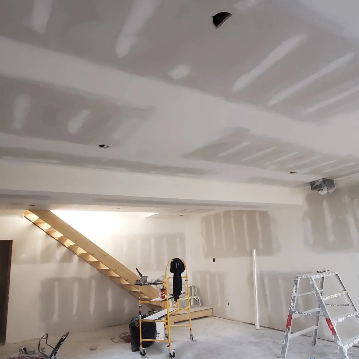 Drywall finishing across an open room, ceiling beam and stair area.